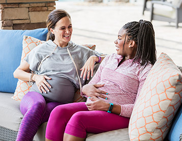 Two pregnant people are sitting together on a sofa. They are both cradling their baby bumps and smiling at each other.
